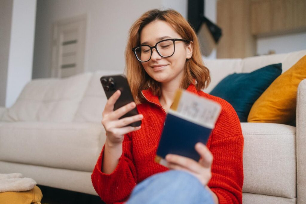 Young woman holding a passport and boarding pass while checking flight details on her smartphone at home.