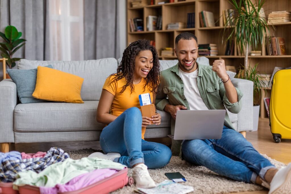Happy couple sitting on the floor booking flights online with a laptop, passports, and luggage packed for travel.