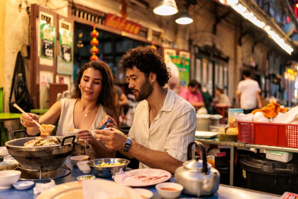 A couple enjoying a meal at a lively night market, sitting at a table with hotpot and various dishes.