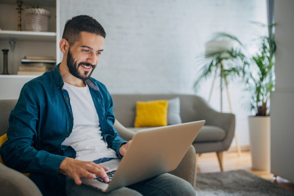 Man smiling while using a laptop at home in a comfortable living room setting.
