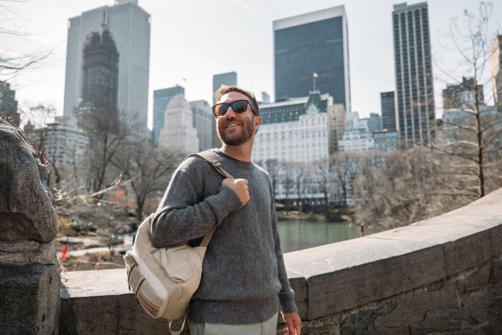 Man smiling with a backpack while sightseeing in Central Park with New York City skyscrapers in the background.