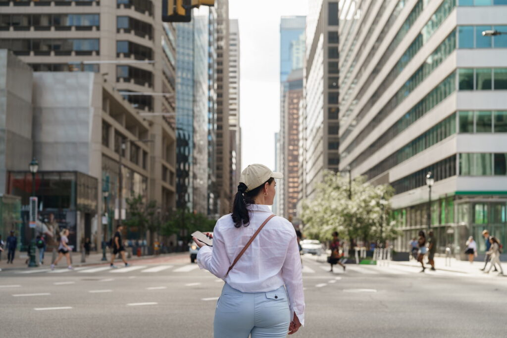 Woman walking through a busy city street holding a phone, surrounded by tall modern buildings.