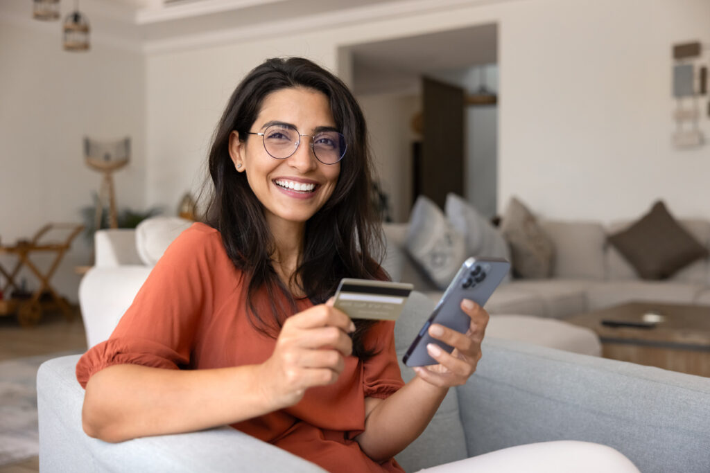 Woman smiling while holding a credit card and smartphone at home.
